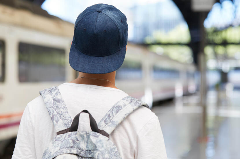 Een jongen met een pet op en een rugzak om loopt op een station, gezien van achteren